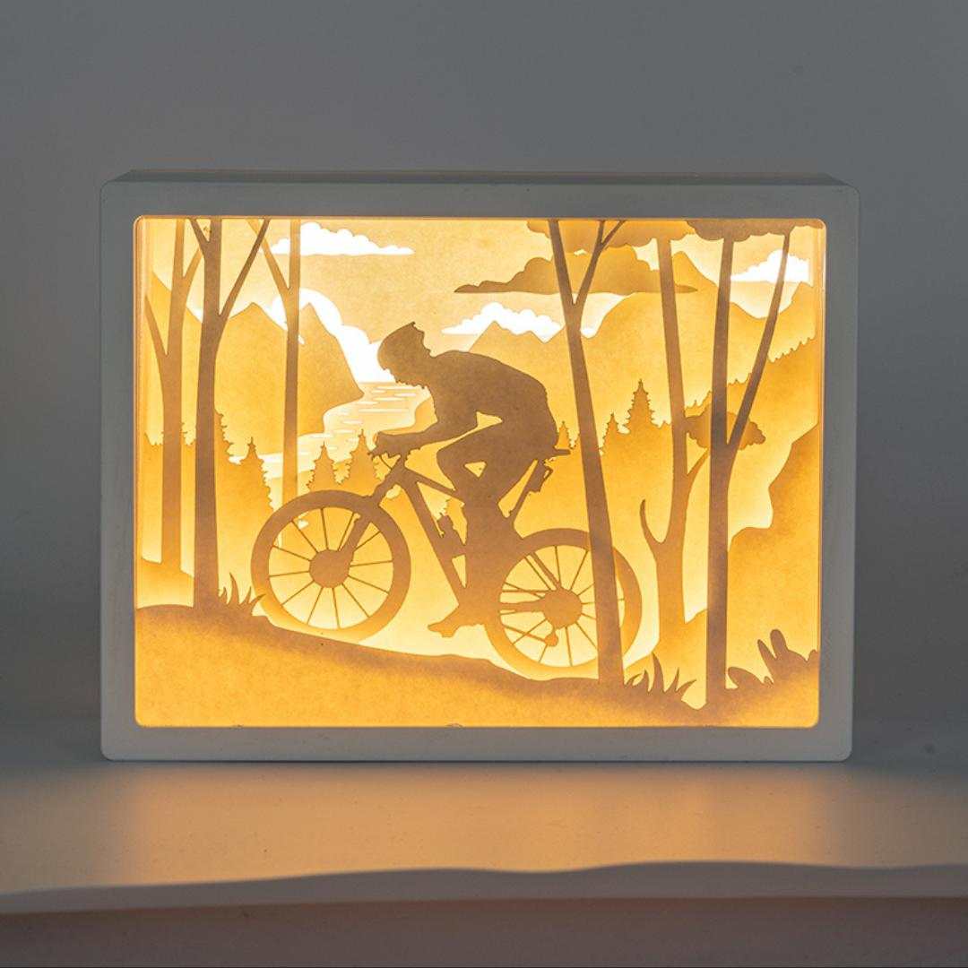 Lámpara 3D Cutout de montaña con ciclista en paisaje forestal iluminado.