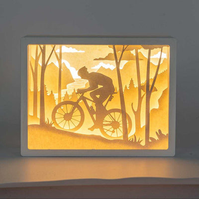 Lámpara 3D Cutout de montaña con ciclista en paisaje forestal iluminado.