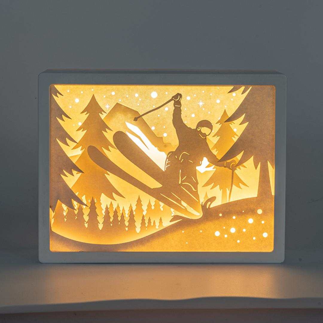 Lámpara 3D Cutout Ski con escena tridimensional de esquí, luz LED cálida, diseño invernal y protección de cristal acrílico.