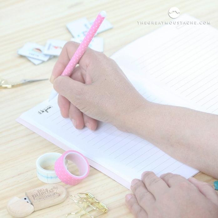 Libreta Grandes Ideas abierta sobre mesa con persona escribiendo, incluye bolígrafo rosa y accesorios de papelería.